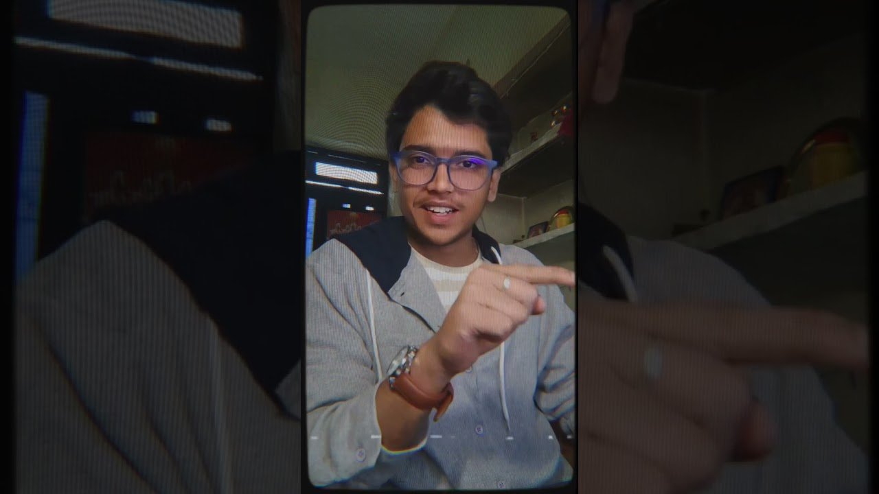 Kunal Kashyap wearing glasses and a casual hoodie, pointing towards the camera with a friendly expression, sitting in a well-lit room with shelves and a window in the background.